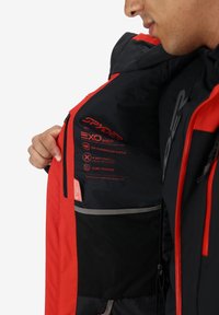 Un uomo con una giacca Spyder rossa e nera apre la fodera interna mostrando le caratteristiche impermeabili, elastiche e termiche stampate all'interno.