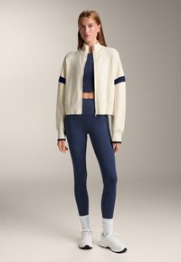 Cremfarbene Bomberjacke mit Reißverschluss und navyblauen Streifen, kombiniert mit einem navyblauen Croptop und passenden Leggings. Das Model trägt weiße Sneakers und strukturierte Socken.