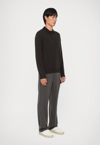 Camisa de polo marrón de manga larga con dobladillo y cuello acanalados, combinada con pantalones grises y zapatillas blancas. Textura suave, diseño ajustado.