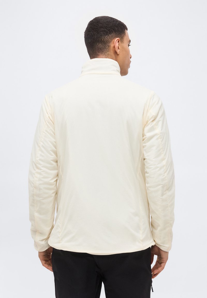 Uomo con giacca color crema dal colletto alto, visto di spalle, in piedi davanti a uno sfondo bianco neutro.