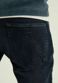 Donkerdenim jeans met een gestructureerde afwerking, uitgerust met schuine achterzakken en contrasterende stiksels. Close-up zicht van de zijkant.