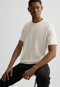 Cremfarbendes T-Shirt aus weichem Stoff mit kurzen Ärmeln und Rundhalsausschnitt. Mit einem dezenten Logo auf der Brust. Trägt dunkle Jeans.