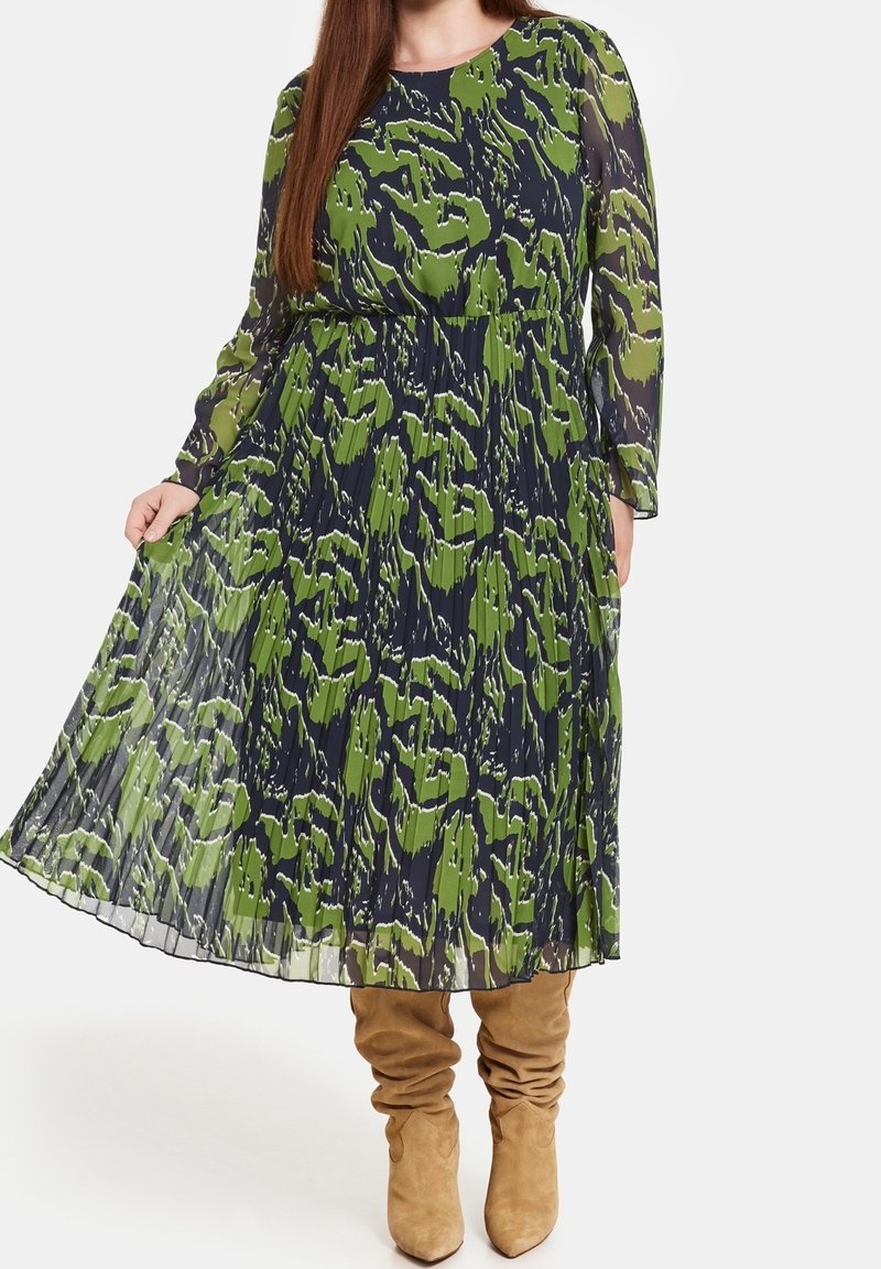 Femme portant une robe plissée à manches longues, à motifs verts et noirs, avec des bottes beiges souples jusqu’aux genoux, debout devant un fond clair.