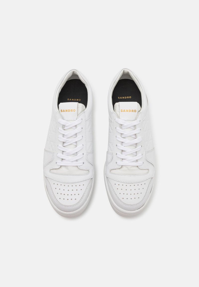 White leather sneakers with perforated details, flat laces, and a cushioned interior. "SANDRO" logo on the tongue in gold.
