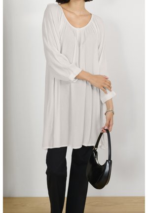 Kobieta ubrana w luźną, białą bluzkę z długim rękawem i czarne spodnie, trzymająca czarną torebkę w jednej ręce, na jednolitym tle.