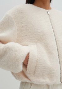 Chaqueta corta color crema hecha de tela texturizada con cierre de cremallera. Cuenta con mangas anchas y una apariencia suave y acogedora.