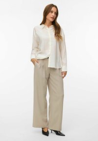 Femme portant un chemisier blanc ample à boutons, un pantalon large beige et des talons noirs à bout pointu, debout avec une main dans la poche.