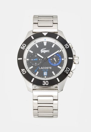 Montre en acier inoxydable avec un cadran noir, des accents bleus et une lunette tournante. Les caractéristiques incluent un affichage double heure et un bracelet en maillons argentés.