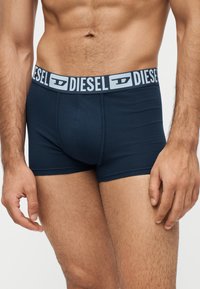 Torse masculin portant un boxer Diesel bleu marine avec une ceinture à logo, montrant l'abdomen, les bras et le haut des cuisses sur un fond uni.