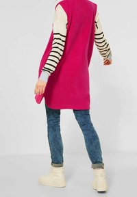 Rosa ärmlös väst med slät textur, bärs över en randig långärmad topp, kombinerad med blå jeans och vita högskochniker.