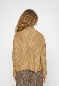 Maglione beige lavorato a maglia con un motivo texturizzato, scollatura rotonda e vestibilità ampia, abbinato a pantaloni a quadri marroni.