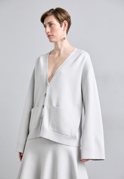 Ανοιχτό γκρί cardigan φτιαγμένο από μαλακό ύφασμα, με βαθύ V-λαιμό, μπροστινά κουμπιά και δύο τσέπες τύπου πατς με φαρδιά μανίκια.