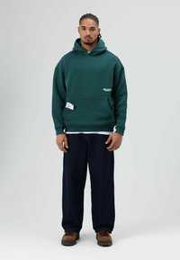 Groen katoenen hoodie met een kangoeroezak, voorzien van een logo op de borst en een label aan de zijkant. Gecombineerd met donkere, wijdvallende broek.