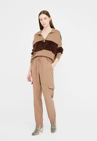 Chándal beige con una franja peluda marrón oscuro, parte superior con cuello, cintura elástica y pantalones estilo cargo; combinado con botas tobilleras con estampado.
