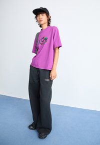 T-shirt oversize violet avec design graphique, assorti à un pantalon de survêtement noir à jambes larges avec le texte "new balance", et des chaussures noires à enfiler.