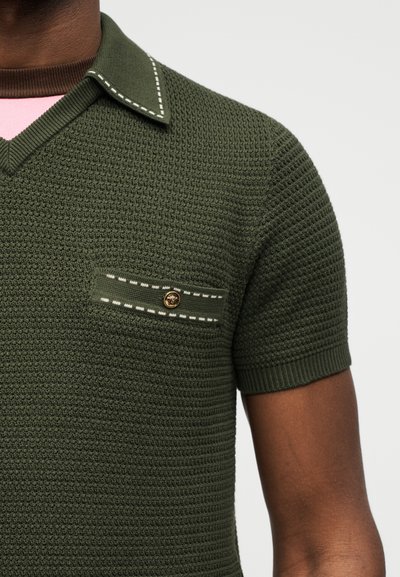 Κοντινή λήψη ενός σκούρου πράσινου πλεκτού πόλο με υφή, μυτερό γιακά, κοντά μανίκια και θήκη στο στήθος με κουμπιά και λευκές ραφές.