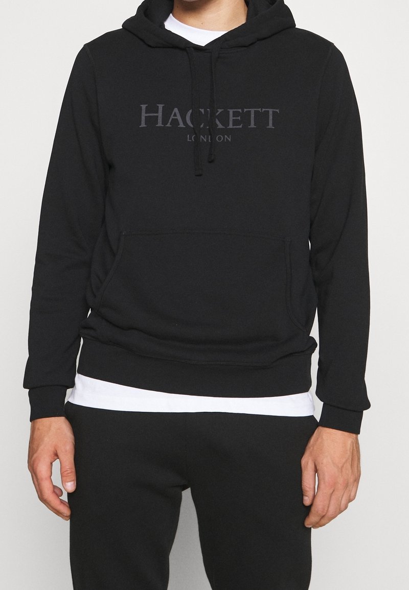 Homme portant un sweat à capuche noir Hackett London par-dessus une chemise blanche et un pantalon noir, debout, les bras relâchés le long du corps.