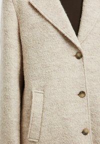 Beige strukturierter Wollmantel mit Reverskragen, drei braunen Knöpfen und einer vertikalen Schlitztasche auf der linken Seite.