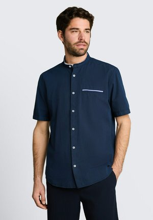 Mann trägt ein marineblaues Kurzarmhemd mit Mandarinkragen und einer hellblau eingefassten Brusttasche.