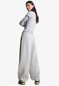 Kvinde med langt mørkt hår iført lagdelte grå toppe, brede lysegrå sweatpants og beige sneakers, stående med armene korslagte og vendt sidelæns.