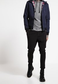 Námorníková bomber bunda s kostkovanou podšívkou, šedá mikina a černé kalhoty se světlým proužkem. Černé šněrovací boty dotvářejí celý outfit.