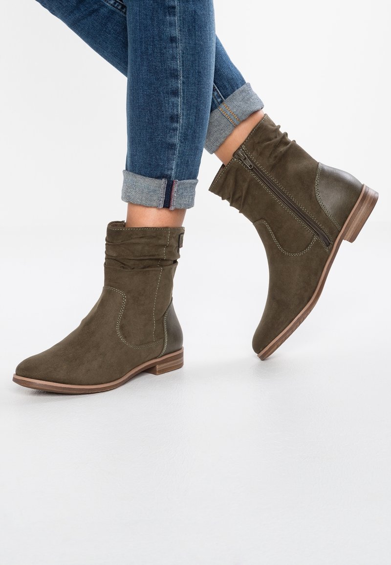 Femme portant un jean bleu retroussé et des bottines en daim vert olive foncé avec fermetures éclair latérales et petits talons en bois.