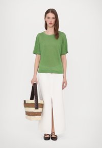 Top verde a maniche corte, gonna bianca con spacco frontale, borsa a tracolla intrecciata con manici marroni e scarpe scure. Design semplice e pulito con materiali testurizzati.
