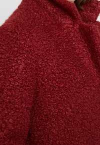 Abrigo de lana rojo con superficie texturizada y esponjosa, cuello alto y un corte extragrande, que muestra un aspecto suave y acogedor.