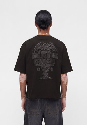 Personne avec les cheveux en chignon porte un t-shirt noir oversize avec un texte graphique blanc délavé dans le dos, associé à un jean ample foncé.
