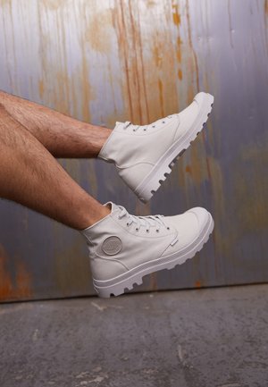 Beine mit weißen High-Top-Leinenschuhen mit dicken Sohlen vor einem rostigen Metallwand-Hintergrund.