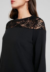 Camisa negra de manga larga con un panel de encaje floral en los hombros. Tejido suave con un cuello redondo y un corte relajado.