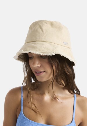 Jonge vrouw met een beige emmerhoed met rafelige rand en een blauw mouwloos topje, kijkt iets naar beneden en glimlacht zacht.