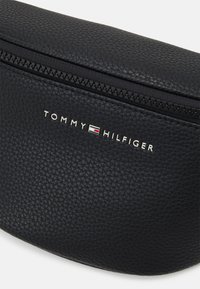 Zwarte leren tas met textuur en een gebogen bovenkant, voorzien van een ritssluiting en een zilveren "TOMMY HILFIGER" logo met rode, witte en blauwe accenten.