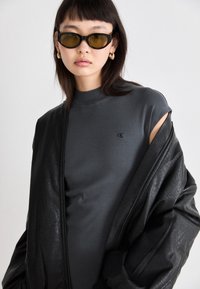 Γκρίζο αμάνικο τοπ με ψηλό λαιμό, που φέρει ένα μικρό λογότυπο. Φορεμένο κάτω από ένα μαύρο δερμάτινο μπουφάν με υφή. Μαύρα γυαλιά ηλίου.