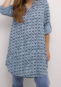 Jasnoniebieska tunika z falistym czarnym wzorem, z zapięciem na guziki i podwiniętymi rękawami. Wykonana z gładkiego materiału o luźnym kroju.