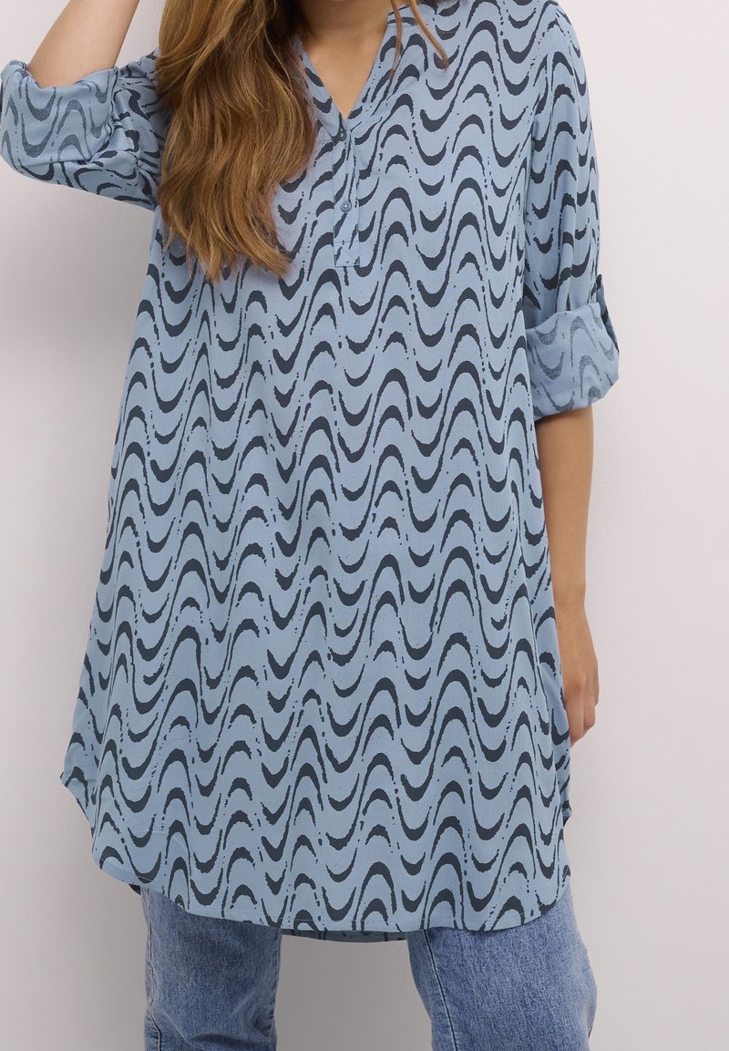 Jasnoniebieska tunika z falistym czarnym wzorem, z zapięciem na guziki i podwiniętymi rękawami. Wykonana z gładkiego materiału o luźnym kroju.