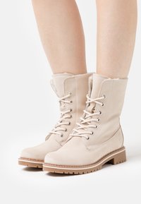 Botas de tobillo de ante beige con cordones, forradas de un material suave, punta redonda y suela de goma texturizada. Presentan ojales metálicos y detalles de costura.