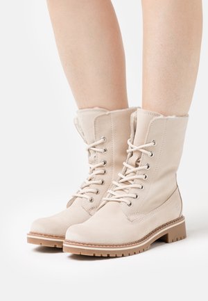 Beige mockasinfärgade snörstövlar i ankellängd med mjukt foder, rund tå och en texturerad gummisula. Har metalldetaljer och sömnadsdekorationer.