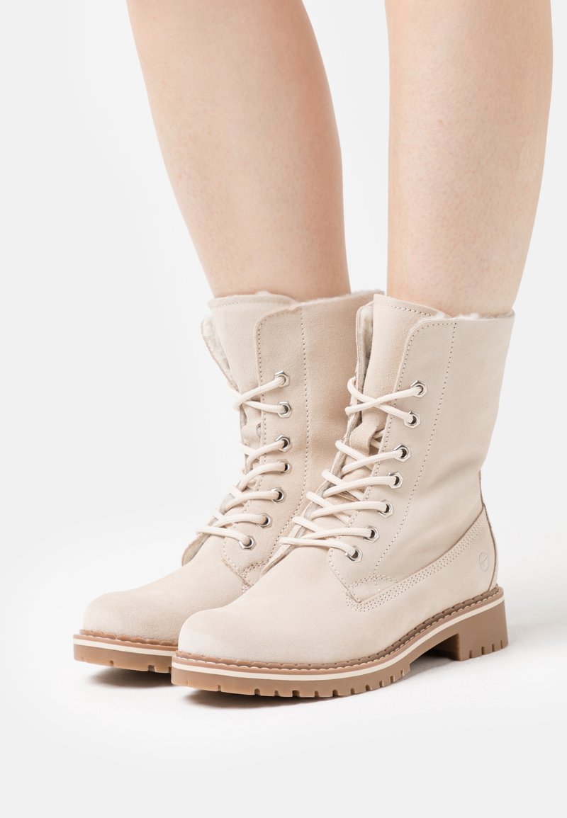 Botas de tobillo de ante beige con cordones, forradas de un material suave, punta redonda y suela de goma texturizada. Presentan ojales metálicos y detalles de costura.
