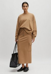 Sweatshirt beige à coupe oversize, associé à une jupe longue assortie avec une poche cargo sur le côté. Sac fourre-tout noir et chaussures.