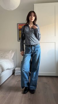 Πουκάμισο με κουμπιά και καρό μοτίβο, μακριά μανίκια, μπλε τζιν με φαρδιά μπατζάκια, μαύρα παπούτσια και μαύρη τσάντα, τοποθετημένα σε ένα φωτεινό δωμάτιο.