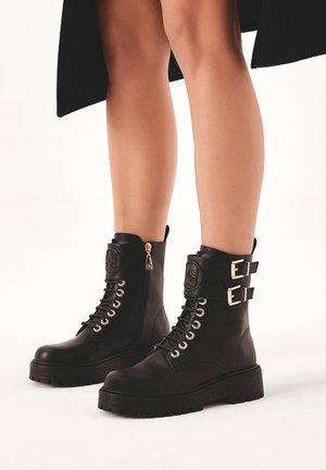 Black leather ankle boots with lace-up design, gold eyelets, dual buckles on the sides, and a decorative zipper pull. Thick, textured sole.
