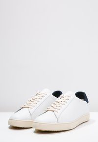 White leather sneakers with a smooth texture, featuring beige laces and a navy heel accent, rounded toe, and textured rubber sole.