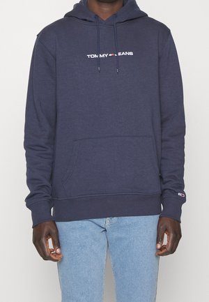 Osoba nosząca granatową bluzę z kapturem Tommy Jeans z przednią kieszenią i jasnoniebieskie jeansy, stojąca na jednolitym tle.