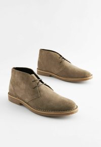 Par de botas desérticas de ante marrón con cordones y suelas cosidas sobre un fondo claro.