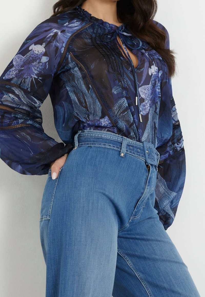 Femme portant un chemisier bleu transparent à motifs floraux avec des manches longues, rentré dans un jean bleu clair taille haute avec une ceinture large.