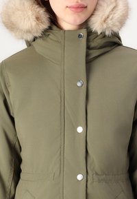 Oliviroheline parka jakk, millel on kunstkarusnahaga kapuuts, hõbedased nööbid ja sile tekstuur. Omab eesmine taskuid ja tikandite detaile.