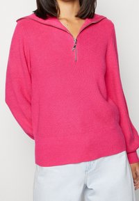 Suéter fucsia con cierre en el cuello, de textura acanalada y mangas largas, combinado con jeans de color azul claro. Presenta un cuello ancho y un ajuste relajado.