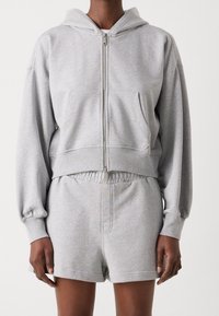 Sudadera gris con cremallera, capucha y bolsillos frontales, combinada con pantalones cortos de chándal a juego. Ambos están hechos de una tela suave y lisa.