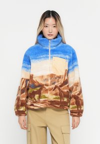Femme portant une veste polaire bleue et marron avec un motif de paysage désertique et de chevaux, une poche poitrine beige et un pantalon cargo beige.